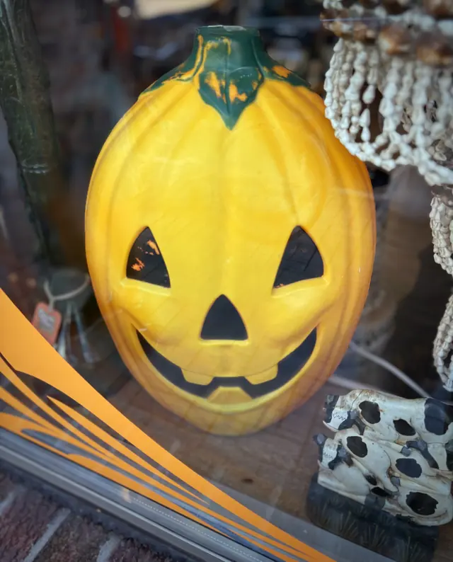 A vintage yellow blow-mould jack-o'-lantern grinning in a shop window, beside a shell curtain and a pair of cow figurines